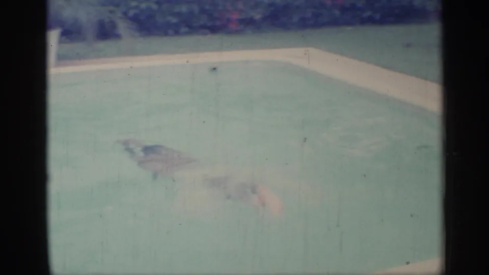 1959 Indiana home-movie frame showing two adults swimming in a backyard-style pool with soft color and visible film grain.