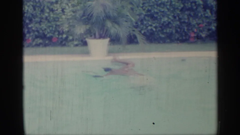 1959 Indiana home-movie frame of an adult swimmer crossing the pool, with a potted palm-like plant and dense hedges behind.