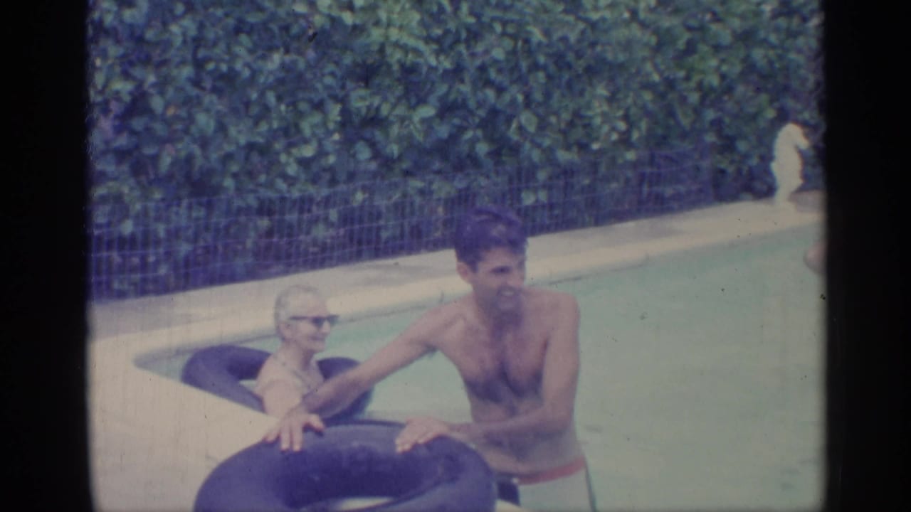 Man in swim trunks and woman in sunglasses lean on inflatable rings in a backyard pool.