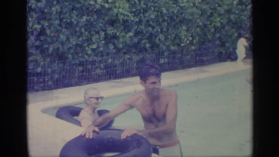 1959 Indiana home-movie frame of adults in a pool, with a man and woman using dark inflatable rings near hedges and fencing.