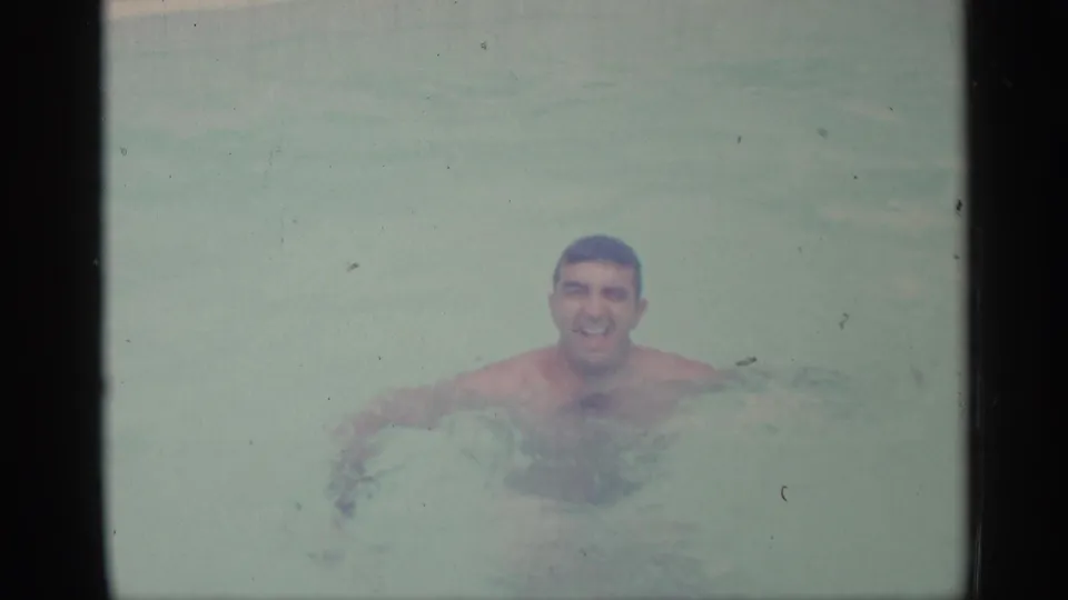 1959 Indiana home-movie frame of a smiling adult man swimming toward the camera in pale outdoor pool water.