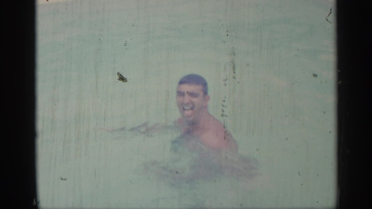 Man in swim trunks smiles while chest-deep in pale pool water.