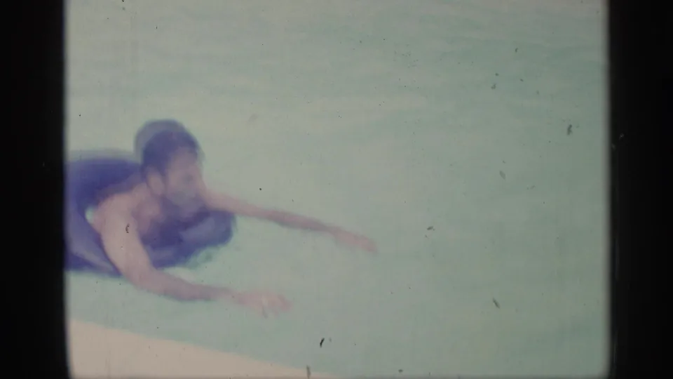 An adult man rests across a dark inner tube while paddling through the backyard pool water.