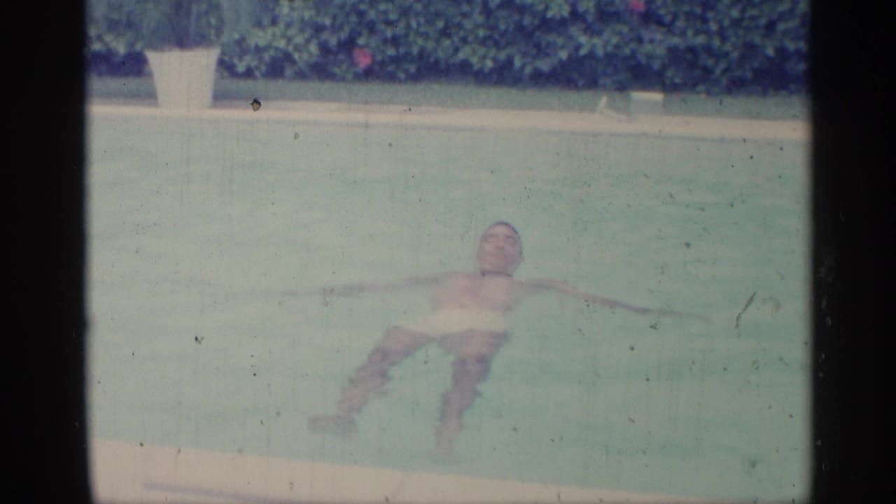 In this 1959 Indiana 8mm frame, a man in white swim trunks floats with arms outstretched in a backyard pool.