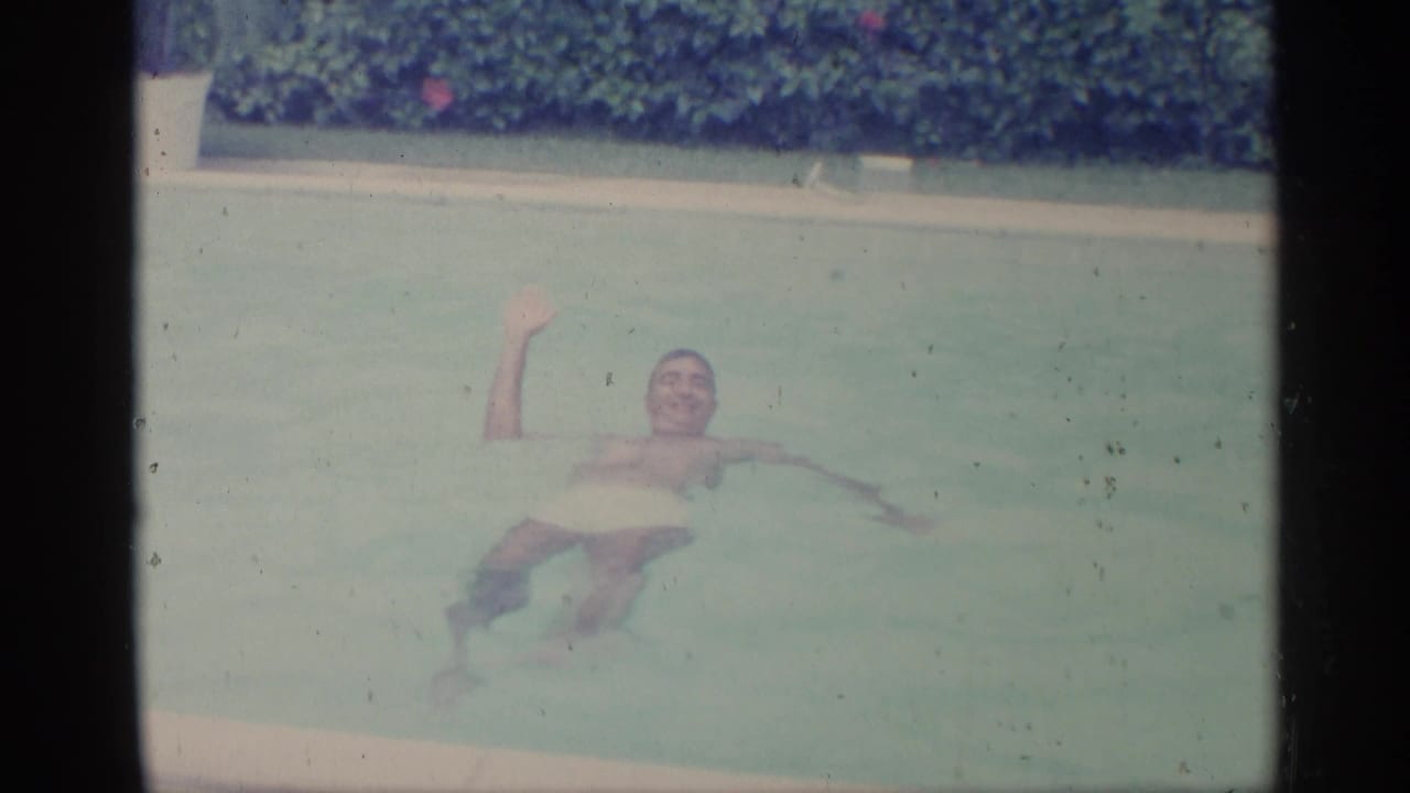 Man in white swim trunks smiling and waving while floating in a pool.