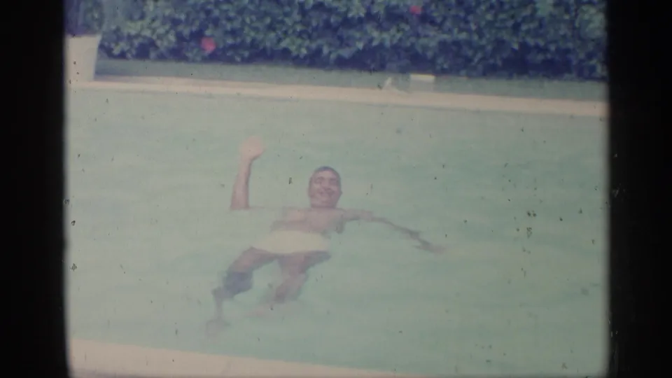 A man in white swim trunks smiles and raises one hand while floating in a pool in 1959 Indiana home-movie footage.