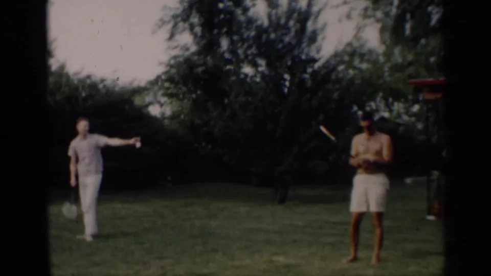 Two adults stand in a grassy Indiana yard as one reaches or throws a flying disc, with trees and shrubs behind them.