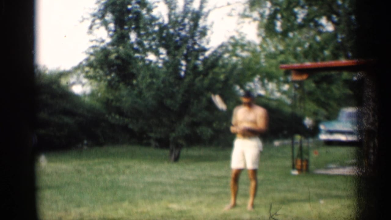 Man in white shorts stands on a lawn holding a badminton racket near trees and a parked car.