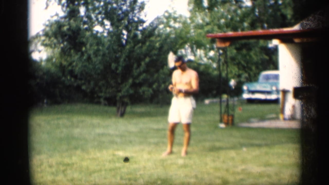 Man with a badminton racket stands barefoot on a lawn near a carport and parked car.