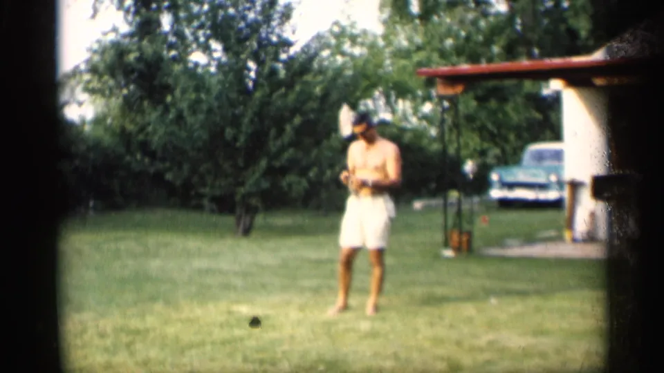 A shirtless man in white shorts stands barefoot with a badminton racket in a 1959 Indiana backyard beside a carport and parked car.
