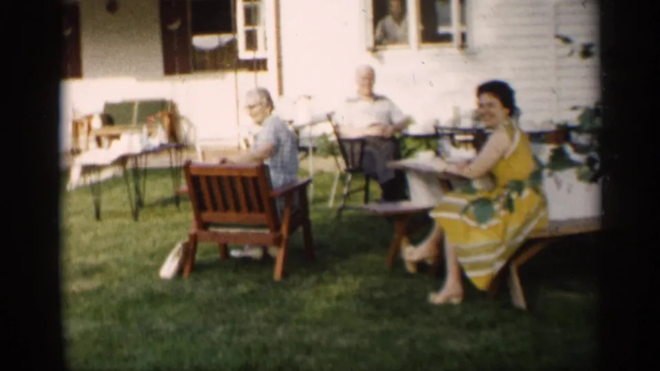 Family members relax in lawn chairs beside a house in Indiana, with a woman in a yellow dress facing the camera.