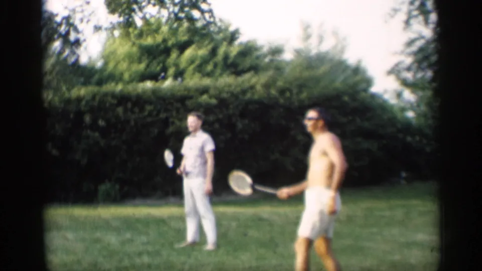 Two men with badminton rackets stand on a 1959 Indiana lawn, one shirtless in white shorts and one in light clothing near tall hedges.