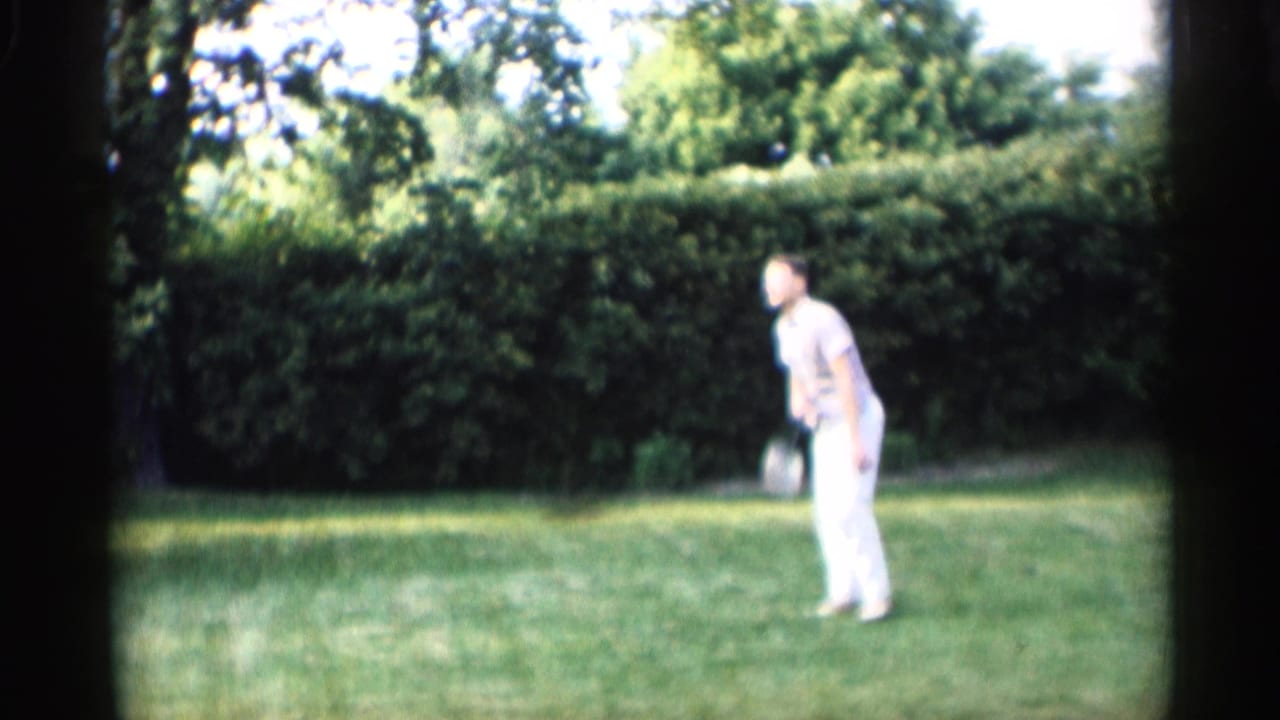 In Indiana 1959 home-movie footage, an adult man in light clothing stands with a croquet mallet on a grassy lawn bordered by hedges.
