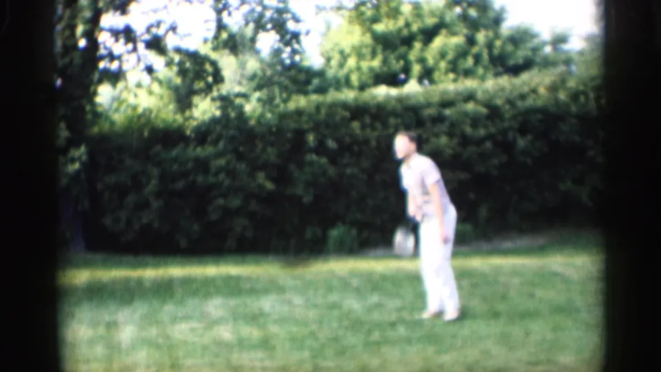 In Indiana 1959 home-movie footage, an adult man in light clothing stands with a croquet mallet on a grassy lawn bordered by hedges.