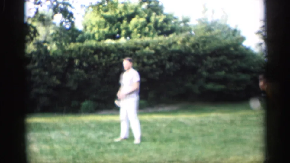 An adult man in white pants holds a croquet mallet on a bright grassy lawn, with dense hedges and trees behind him.