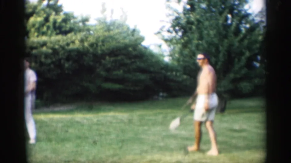 A shirtless adult man in white shorts and sunglasses crosses the grassy Indiana lawn with a croquet mallet in 1959 home-movie footage.