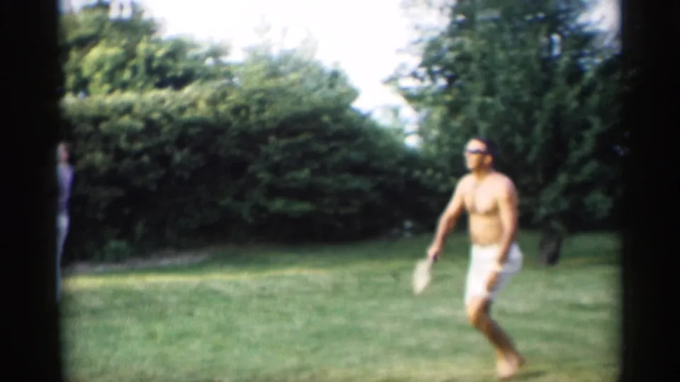 A shirtless adult man in sunglasses prepares or moves during a croquet turn on a sunny lawn bordered by shrubs and trees.
