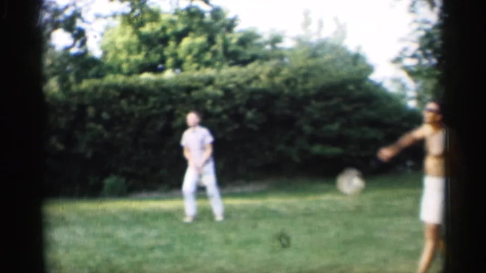 In 1959 Indiana home-movie footage, two men play badminton on a backyard lawn bordered by hedges.