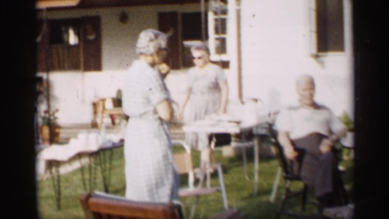 Adults gather beside outdoor tables and chairs near a house.