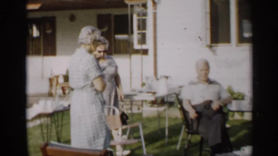 Two women in dresses stand near lawn chairs while an older man sits outside a house in Indiana home-movie footage.