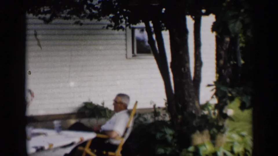 An older man relaxes in a lawn chair under a tree beside a house during a 1959 Indiana backyard scene.