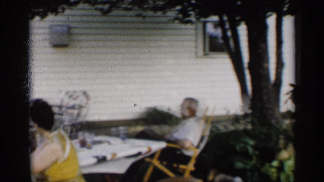 Adults seated at backyard tables near a white-sided house and trees.