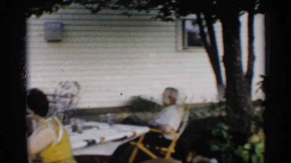 1959 Indiana home-movie frame showing adults relaxing at outdoor tables in a shaded backyard beside a white-sided house.