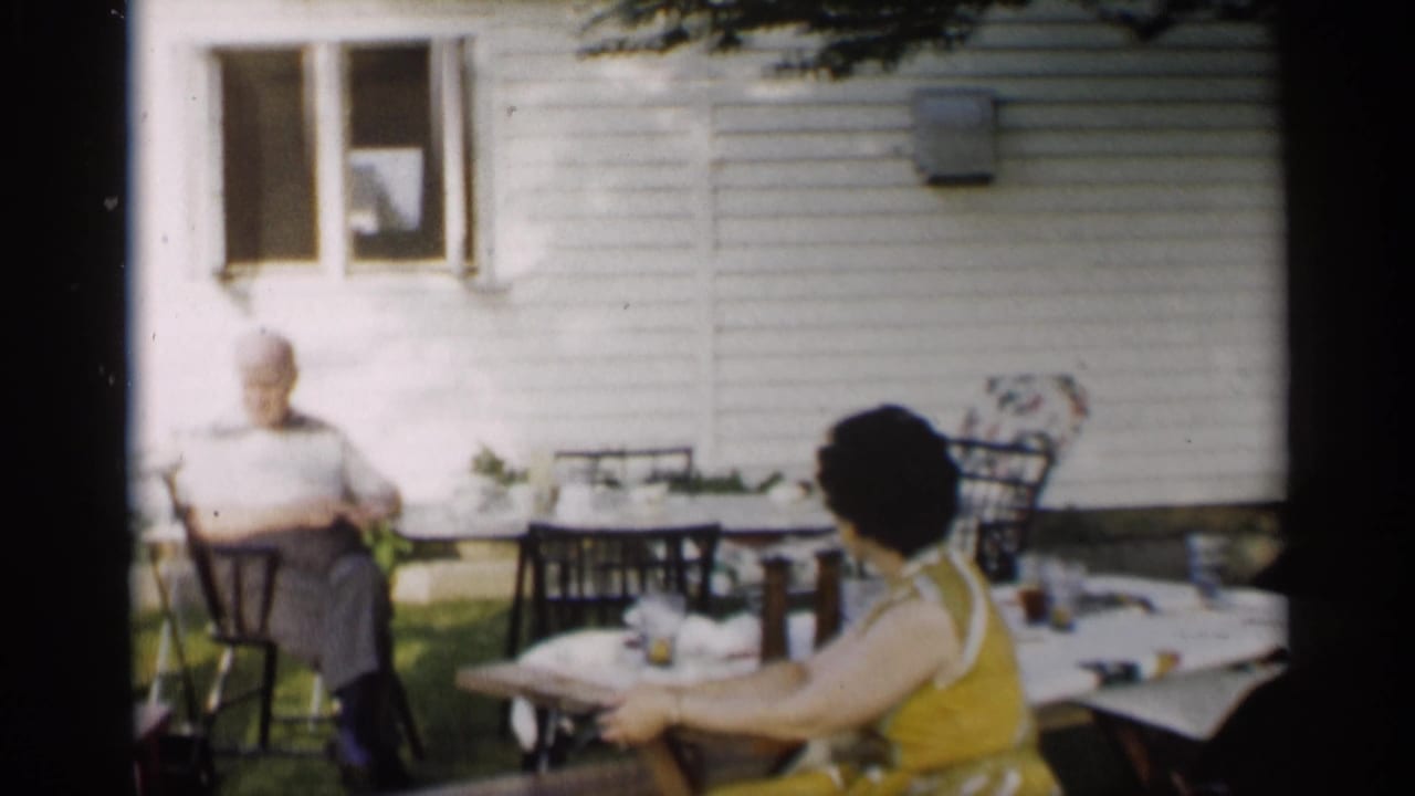 Elderly man seated in a chair while a woman sits at a table nearby.