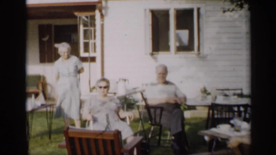 1959 Indiana home-movie frame showing older adults seated and standing in a backyard beside an open window and patio furniture.