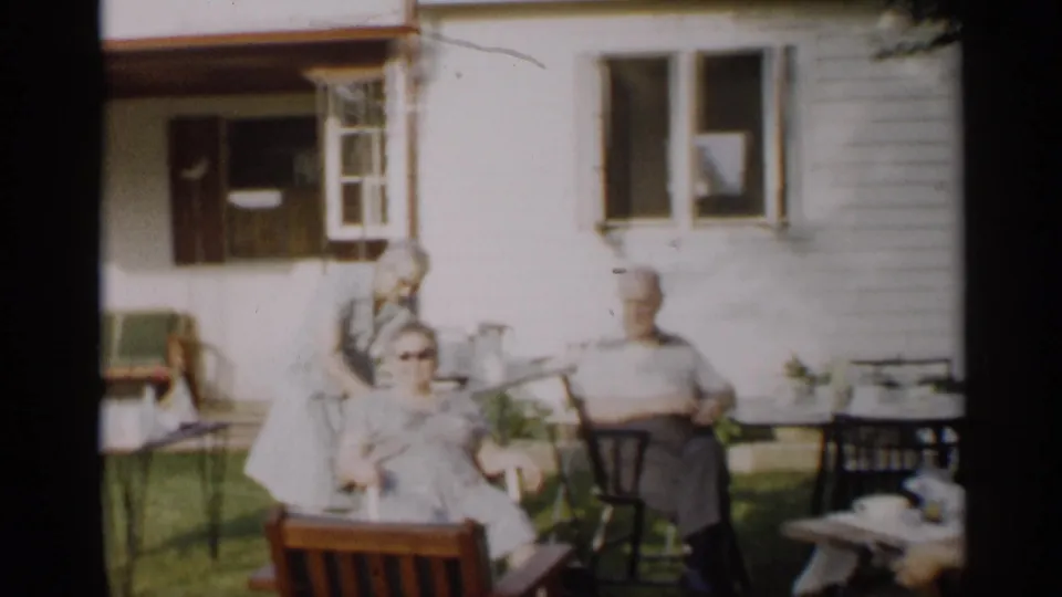 1959 Indiana backyard frame with older adults seated near a white-sided house, lawn furniture, and an open window.