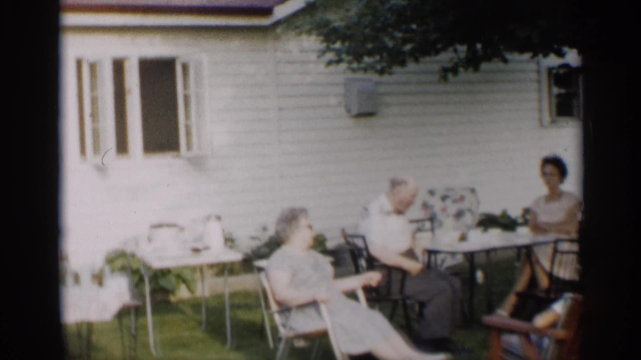 Adults sit around a small backyard table beside a white house on a sunny lawn.
