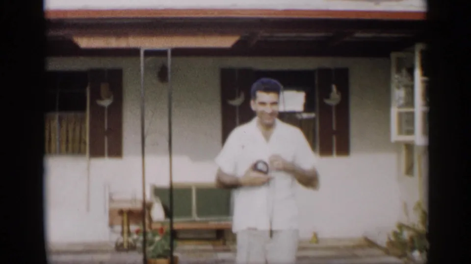 The man pauses near the porch of the white-sided home, facing the camera with windows and porch furniture behind him.