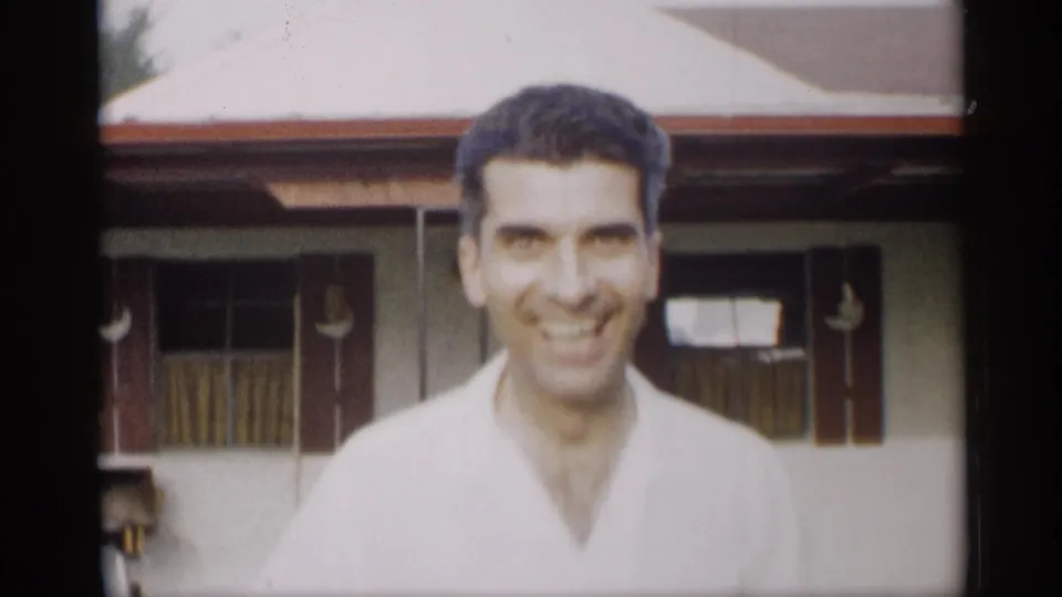 Close-up of the smiling young man, with the porch windows and white house exterior softly visible behind him.