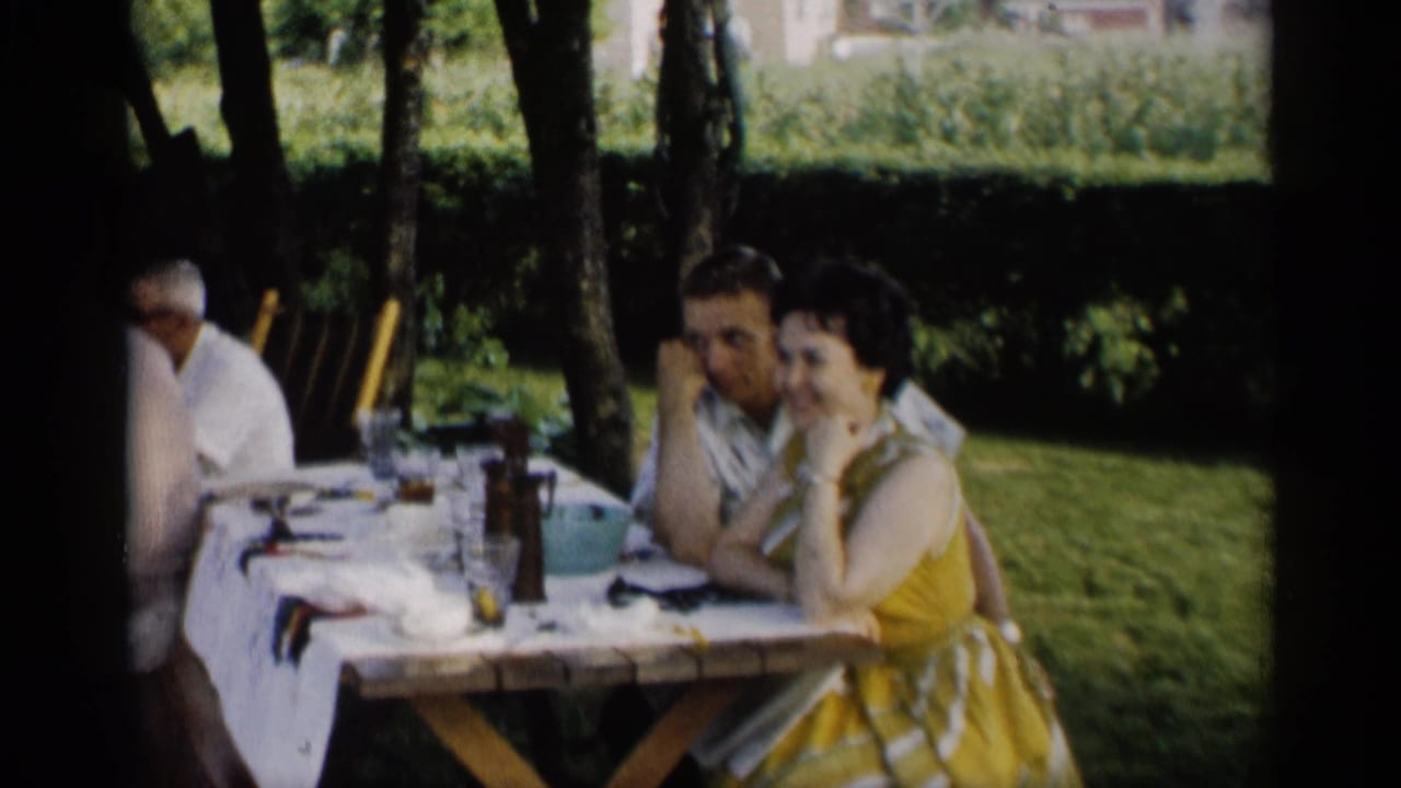 1959 Indiana home-movie frame of adults at an outdoor picnic table, with a woman in a yellow patterned dress facing the camera.