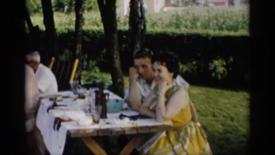 1959 Indiana home-movie frame of adults at an outdoor picnic table, with a woman in a yellow patterned dress facing the camera.