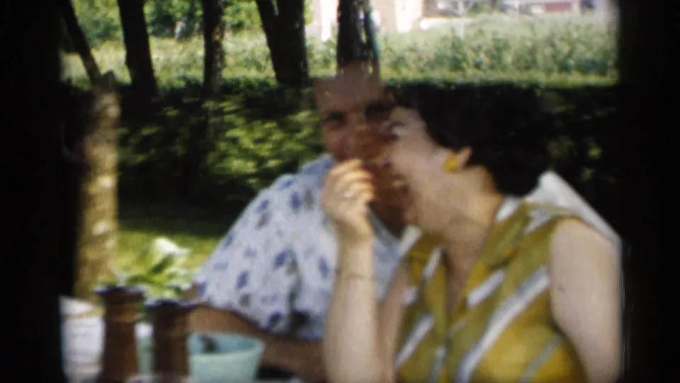 Close 1959 Indiana home-movie view of a woman in a yellow patterned dress at a picnic table, with another adult behind her.