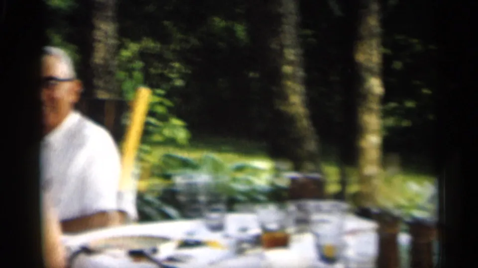 1959 Indiana home-movie frame showing an older man in glasses beside a picnic table with cups, glasses, and serving items under trees.