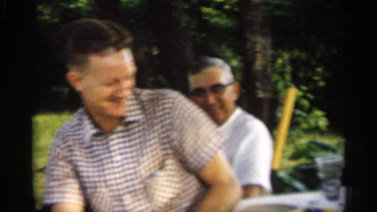 Two men seated outdoors at a backyard table, one laughing in a checkered shirt.