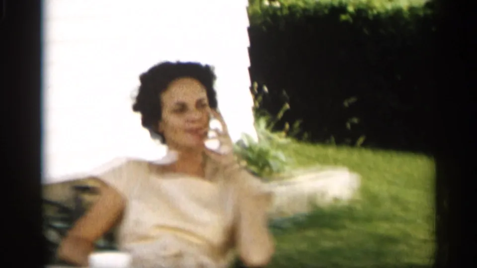 A woman in a light dress sits in a sunny Indiana backyard, holding a corded telephone receiver beside grass and hedges.