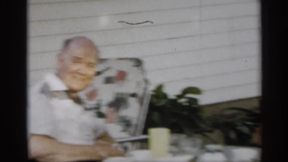 An older man sits at an outdoor table near white house siding, with a cup, glassware, plants, and lawn furniture visible.