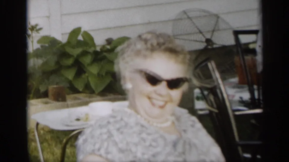 A smiling woman wearing dark sunglasses sits in a backyard near white siding, potted plants, a small table, and lawn chairs.