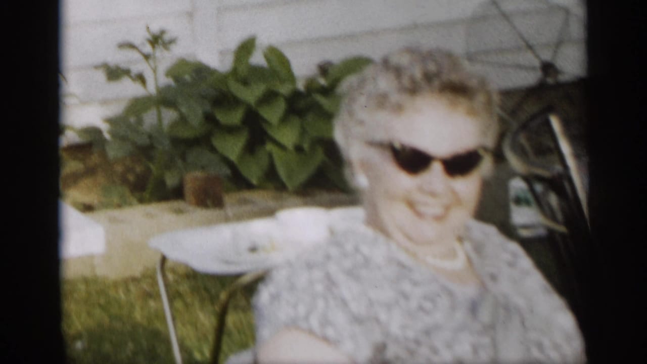 Elderly woman in sunglasses sits outdoors near lawn furniture and plants.