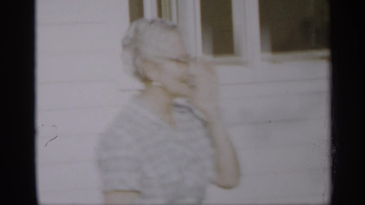 Elderly woman stands near a white house, blurred by motion.