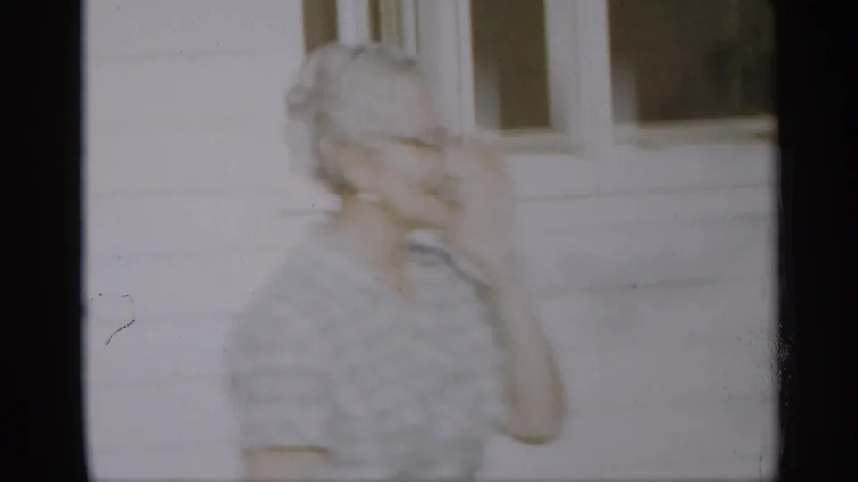 An elderly woman appears near a white house exterior in a soft, motion-blurred Indiana 1959 home-movie frame.