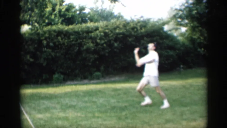 A person plays badminton on a green backyard lawn, with hedges and trees behind them in Indiana 1959 home-movie footage.