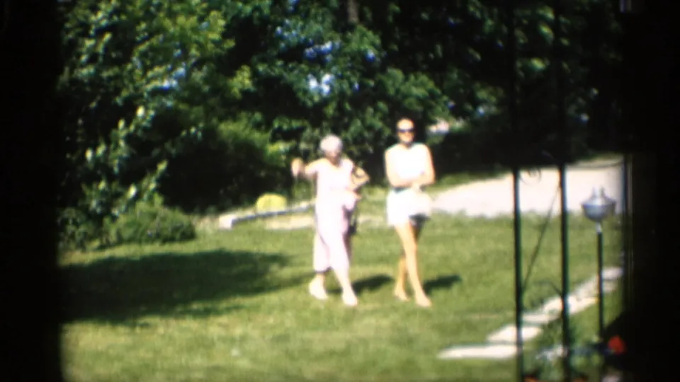 In 1959 Indiana home-movie footage, two women walk across a green lawn toward a house, with trees and a path behind them.
