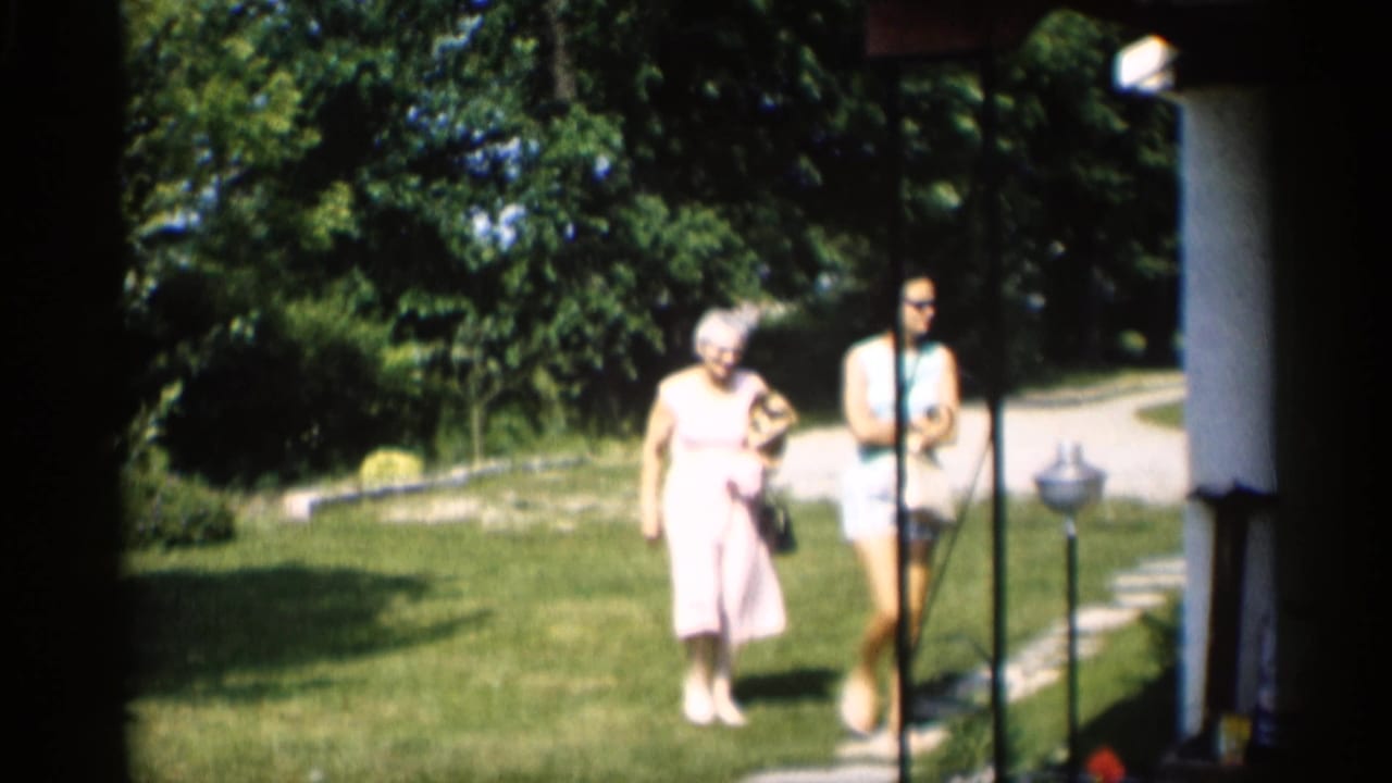 An older woman in a light dress carries a small dog while a younger woman walks beside her across an Indiana lawn in 1959 footage.