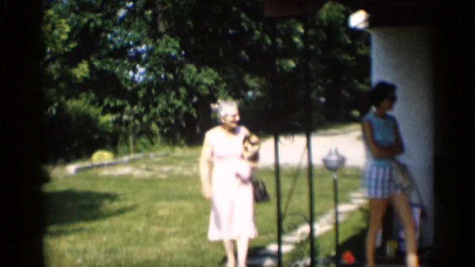 A woman carrying a small dog approaches the porch area as another person stands in shadow near the house exterior.