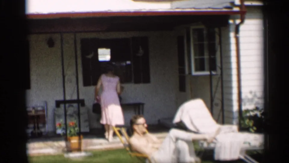 At the house porch, a woman in a light dress stands near the doorway while a man reclines in a lawn chair on the grass.