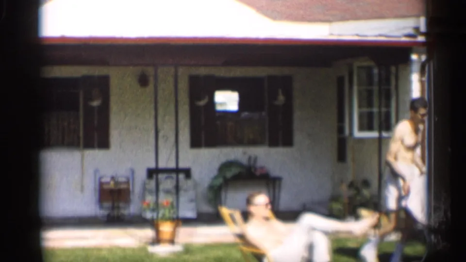A backyard view shows a reclining man on the lawn and another adult near the side of a white Indiana house in 1959 footage.